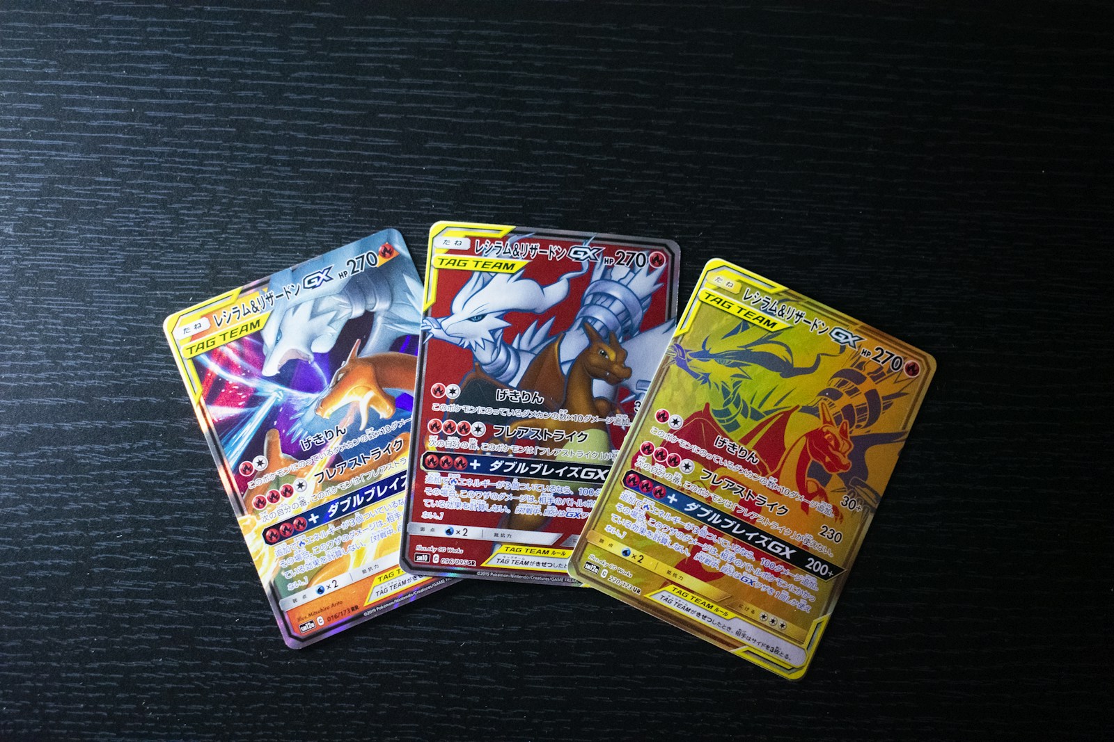 Tres cartas japonesas de Reshiram y Charizard GX con ilustraciones diferentes sobre una mesa oscura.