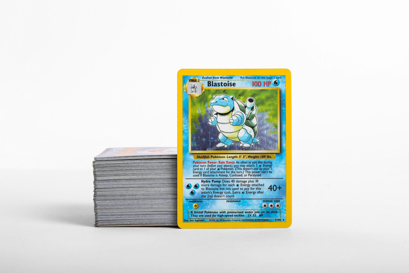 Una carta Pokémon de Blastoise junto a una pila de cartas coleccionables sobre una superficie clara.